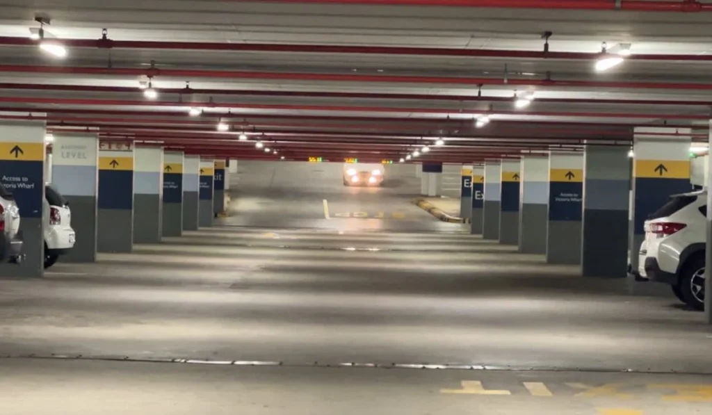 Indoor parking garage with rows of pillars, overhead red pipes, and cars parked along the sides